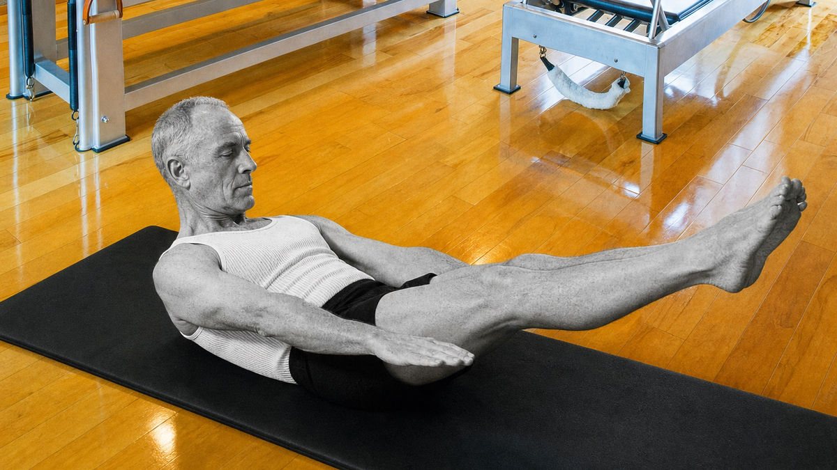 Joseph Pilates demonstrating mat work, legs extended and spine lifted, in the West Hollywood Pilates studio with the Cadillac and reformer behind him
